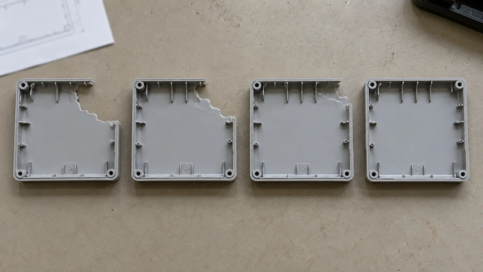 Overhead view of four matching pale plastic molding trial samples showing progressively more complete filling of a ribbed enclosure cover